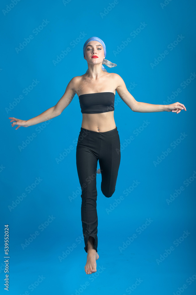 Sport, fashion and make-up concept. Slim and beautiful woman with black and tight sport outfit, scarf and red lipstick jumping in air. Graceful pose freeze in motion. Blue studio background
