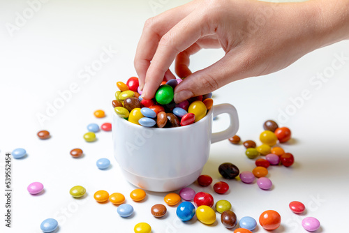 Fotografija Taza blanco con bolas de chocolate de colores con fondo blanco y mano cogiendo g
