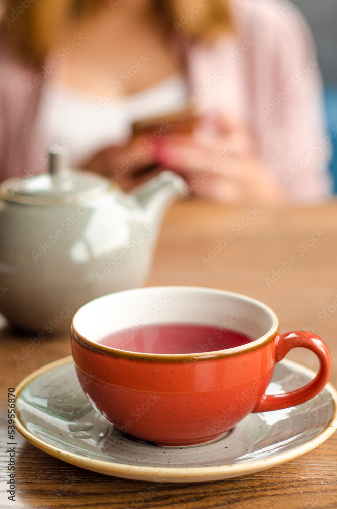 Red cup of red tea, a gray teapot stand on a table in a cafe. In the background, a young woman is working on her smartphone Red cup of red tea, a gray teapot stand on a table in a cafe. In the background, a young woman is working on her smartphone