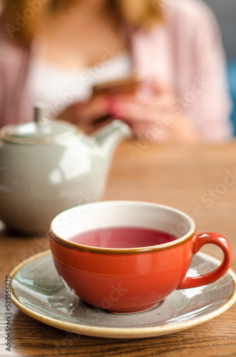 Red cup of red tea, a gray teapot stand on a table in a cafe. In the background, a young woman is working on her smartphone