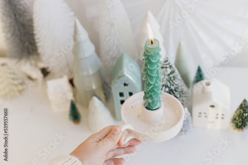 Merry Christmas! Hand holding stylish christmas tree candle on background of miniature winter village on white table. Modern little Christmas trees and houses. Holiday advent