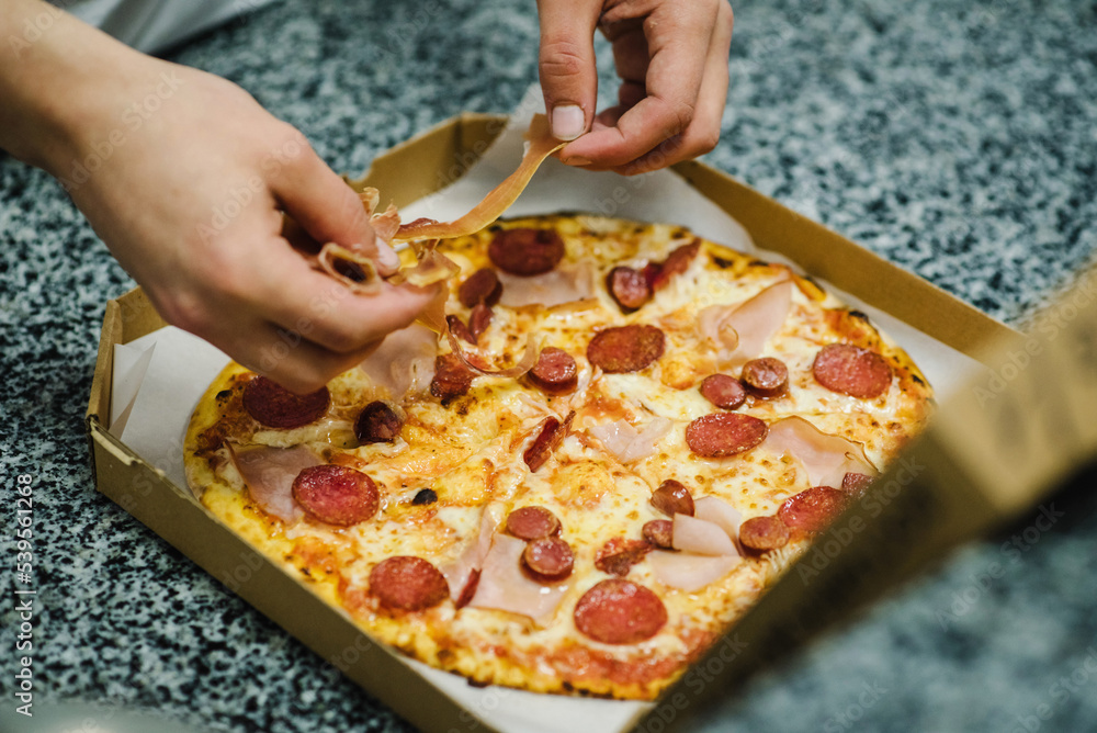 Baked pizza in a box. Traditional Italian pizza with meat, salami, cheese. Chef puts the prosciutto. Food delivery. The last process before pizza is served. Self-pickup or home delivery. Top view.