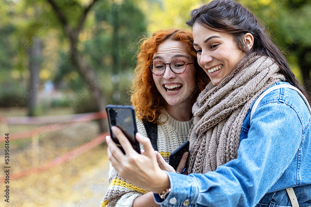 Two student woman friends in urban public park use mobile phone and smile Two student woman friends in urban public park use mobile phone and smile
