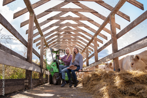 Portrait of two farmers women posing on farm vehicle around hay in barn.