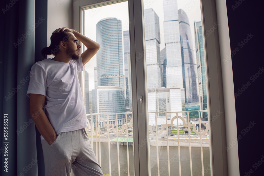 man with long hair, beard and glasses stands at a large window in an apartment with a view of a skyscraper in the morning. the guy is dressed in a white T-shirt and sweatpants barefoot. man with long hair, beard and glasses stands at a large window in an apartment with a view of a skyscraper in the morning. the guy is dressed in a white T-shirt and sweatpants barefoot.