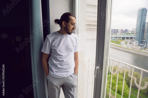 man with long hair, beard and glasses stands at a large window in an apartment with a view of a skyscraper in the morning. the guy is dressed in a white T-shirt and sweatpants barefoot.