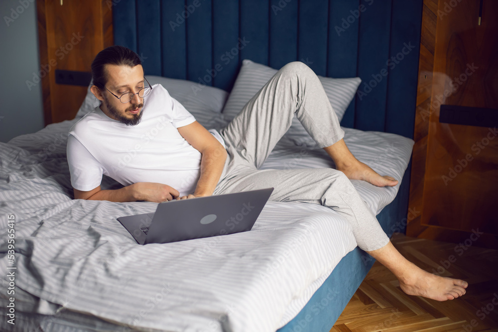 man in a white T-shirt, a freelancer with glasses, sits on the bed at home with a laptop and works remotely in the morning. man in a white T-shirt, a freelancer with glasses, sits on the bed at home with a laptop and works remotely in the morning.