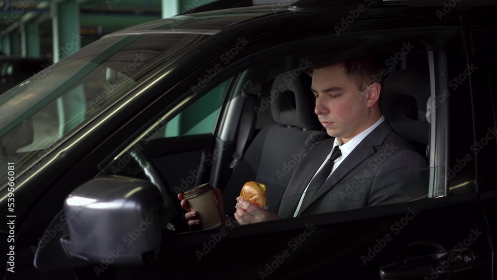 A young businessman is having lunch in a car in a parking lot. A man in a suit eats fast food and enjoys the taste. The businessman eats a cheeseburger and drinks coffee.