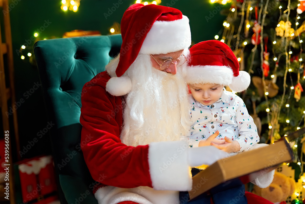 Santa Clause reads fairytale to a little boy while sitting near Christmas tree. New Year concept Santa Clause reads fairytale to a little boy while sitting near Christmas tree. New Year concept
