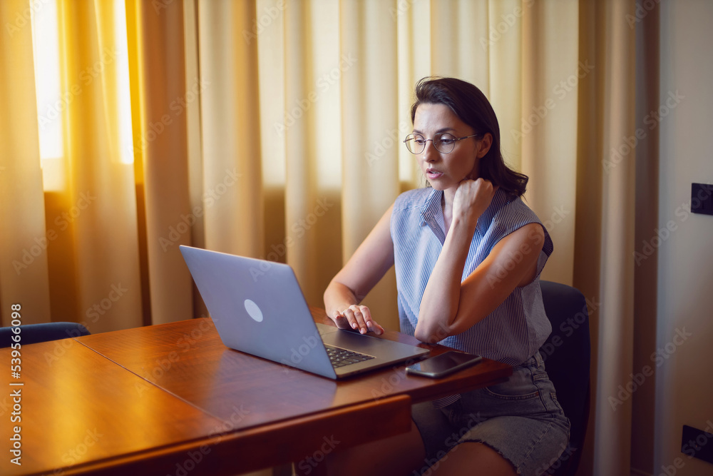 portrait of a woman with glasses a freelancer with a laptop sitting at a table in an apartment. portrait of a woman with glasses a freelancer with a laptop sitting at a table in an apartment.
