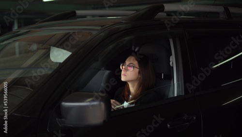 A business woman looks in a car mirror and straightens her hair. A young woman in glasses and a suit in a parking lot sits in a car and straightens her hair.