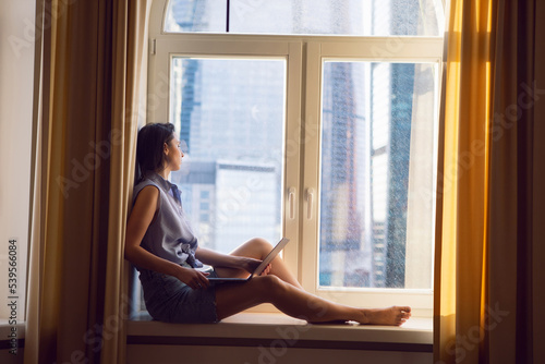 business woman freelancer is sitting on the windowsill by the window with a laptop and working.