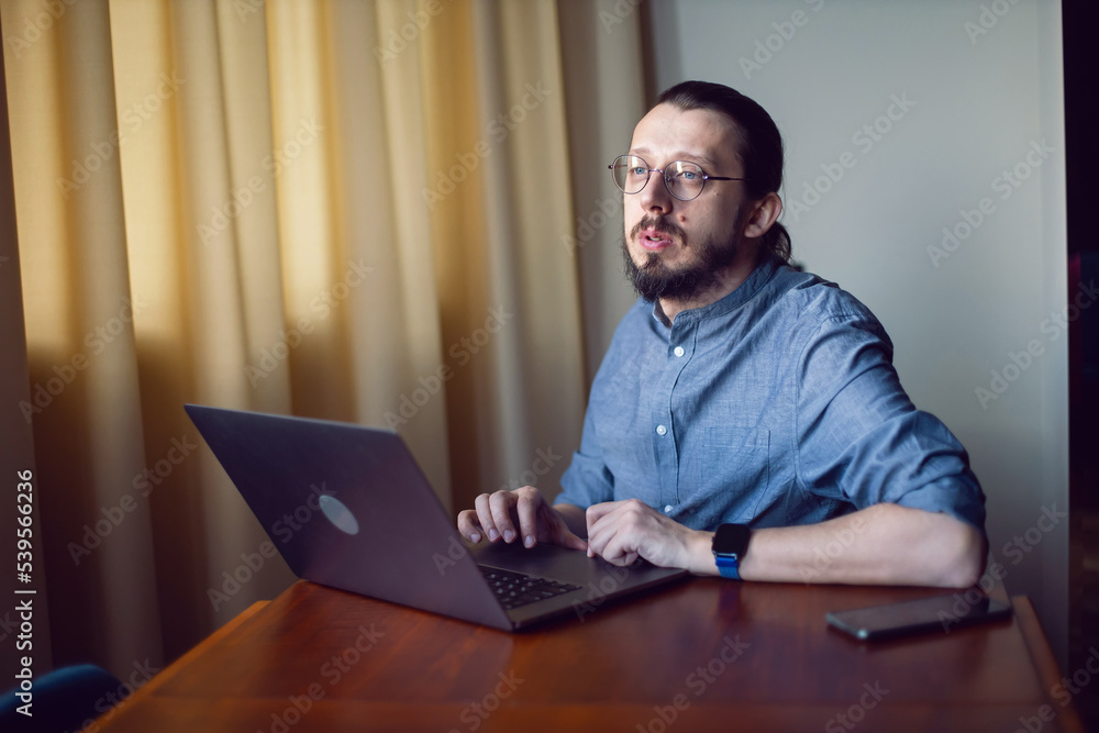 portrait of a businessman a man with a beard and glasses is sitting at a laptop at a wooden table at home. the freelancer works remotely next to the window the light is natural. portrait of a businessman a man with a beard and glasses is sitting at a laptop at a wooden table at home. the freelancer works remotely next to the window the light is natural.