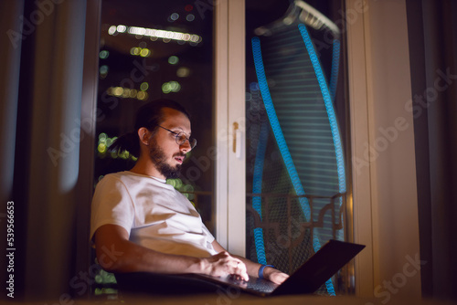 Free Download freelance man in glasses working in shorts and a T-shirt sits at the window on the floor in front of a skyscraper at night. - GFXLoad