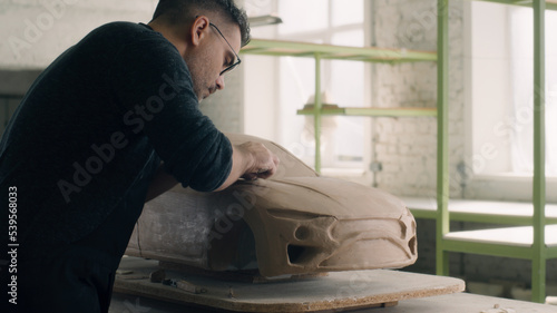 Senior car designer sculpting a electric car model prototype. Uses wired loop tool to carve out chunks of clay for the design details and smooth surface. Automotive industry.