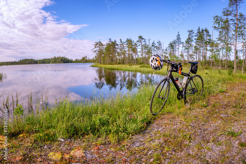 Modern carbon road racing bike stands close to forest lake in front of rare pine trees. Summer Sunny day. Standalone black bike, white helmet, two water sport bottles mounted on frame