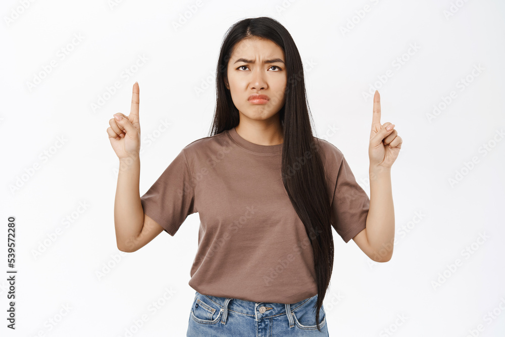 Disappointed asian woman pointing, looking up with sad, regret face, showing smth bad, standing over white background Disappointed asian woman pointing, looking up with sad, regret face, showing smth bad, standing over white background