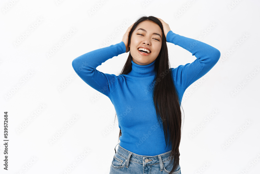 Portrait of beautiful asian woman with long, natural dark hair, touching her hairstyle, laughing and smiling, standing over white background Portrait of beautiful asian woman with long, natural dark hair, touching her hairstyle, laughing and smiling, standing over white background