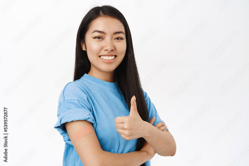 Covid-19, pandemic and healthcare. Smiling korean woman with band aid on shoulder, promotes vaccination, shows thumbs up, coronavirus vaccine, stands over white background Covid-19, pandemic and healthcare. Smiling korean woman with band aid on shoulder, promotes vaccination, shows thumbs up, coronavirus vaccine, stands over white background