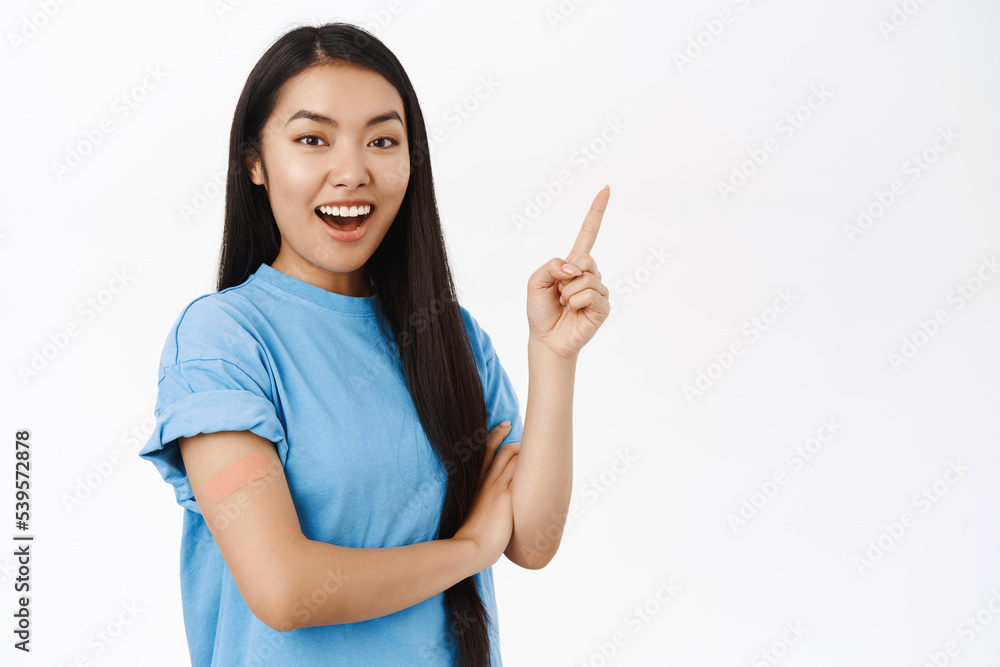 Enthusiastic asian girl got vaccinated, has band aid patch on shoulder after covid vaccine, smiling and pointing at copy space banner, showing coronavirus info announcement Enthusiastic asian girl got vaccinated, has band aid patch on shoulder after covid vaccine, smiling and pointing at copy space banner, showing coronavirus info announcement