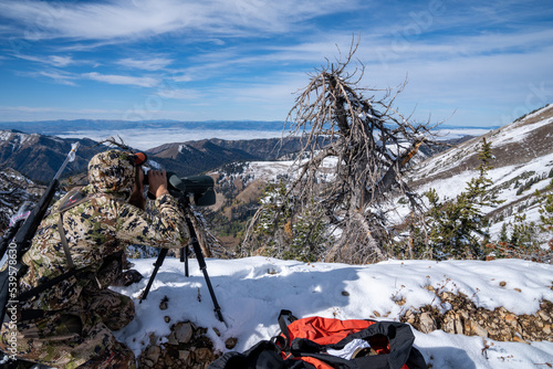 Hunter wearing camo uses a spotting scope to view deer during a hunt in the snow