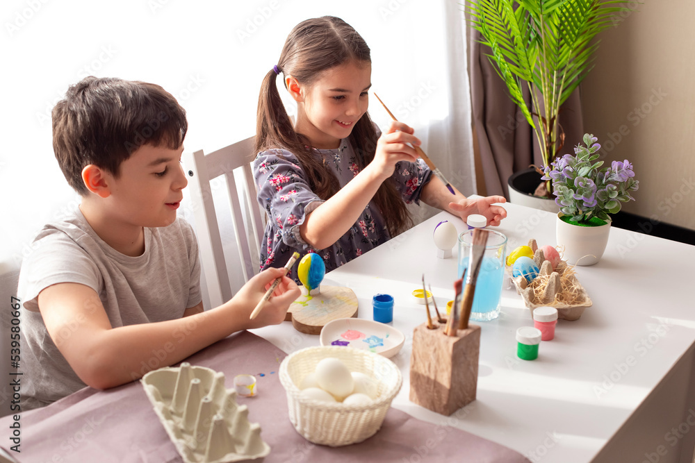 A happy brunette little girl and boy sits at a white table near the window, paints eggs A happy brunette little girl and boy sits at a white table near the window, paints eggs