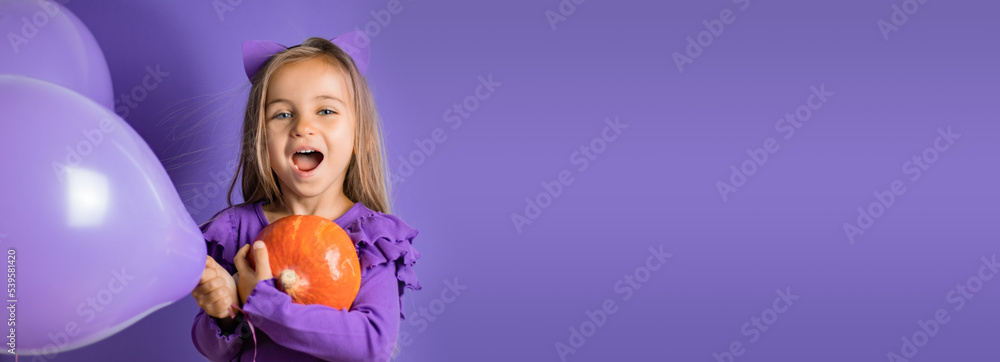Fototapeta premium Banner Gorgeous little girl with cat ears, balloons and a pumpkin in her hand on a purple background. Halloween concept