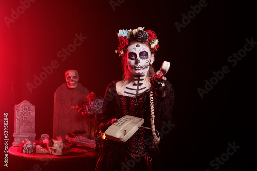 Creepy goddess model using landline phone to answer call, holding office cord telephone on mexican traditional holiday. Looking like la cavalera catrina on holiday, celebrating dios de los muertos.