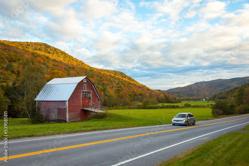 Minivan car driving on scenic Vermont road passing red barn and fall foliage mountains during autumn  © Lenspiration