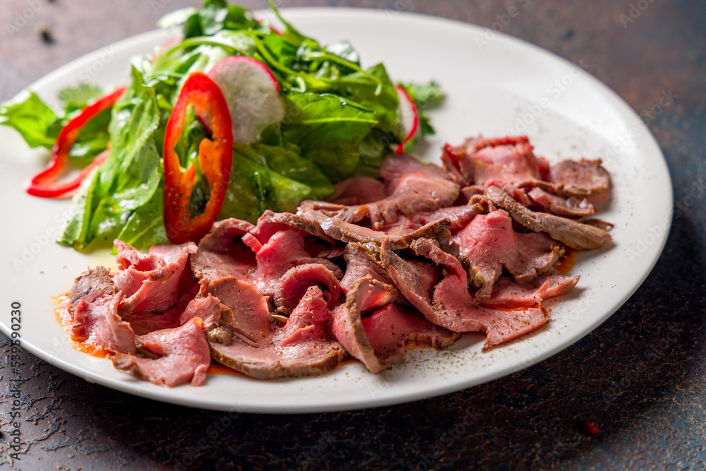 Sliced roast beef with green salad on white plate on dark stone table macro close up Sliced roast beef with green salad on white plate on dark stone table macro close up