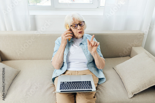 a cute elderly woman is sitting at home working on a laptop resting on the couch and talking on the phone actively gesturing with her hand