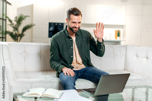 Distant conversation. Positive caucasian man, freelancer, sits on a sofa in living room, uses his laptop, talking online by video conference with colleagues or clients, makes greeting gesture, smiles