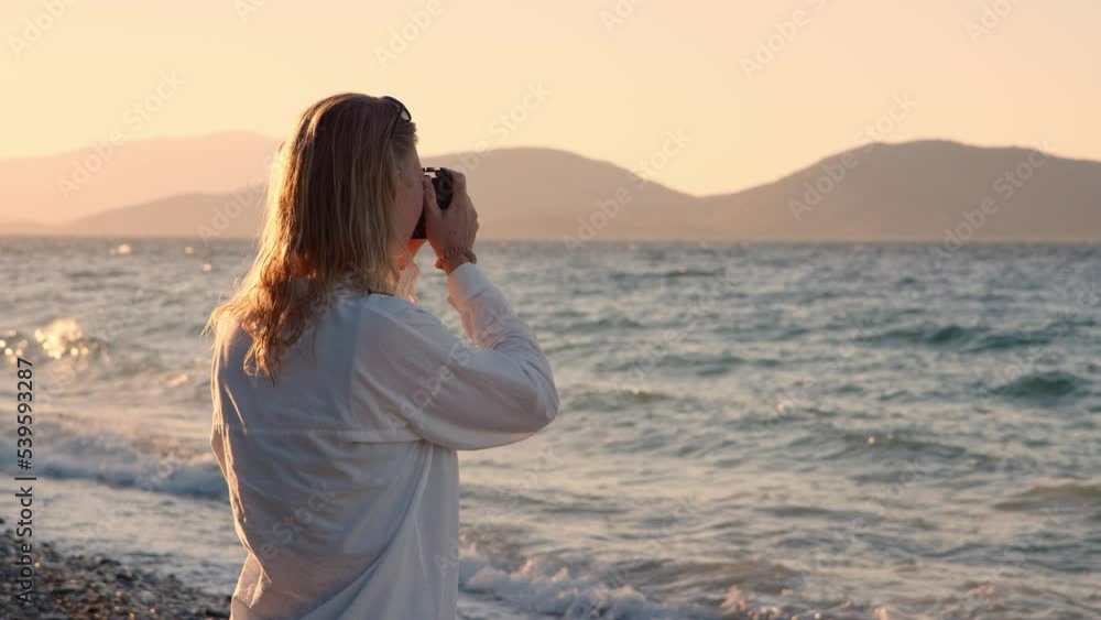 Professional photographer blonde young woman near sea on shore. sunset, unrecognizable girl photographs nature and ocean waves at dawn. traveler captures moments of nature while traveling. Professional photographer blonde young woman near sea on shore. sunset, unrecognizable girl photographs nature and ocean waves at dawn. traveler captures moments of nature while traveling.