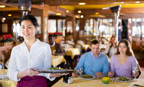Polite smiling asian waitress standing with serving tray  inviting to modern cozy restaurant