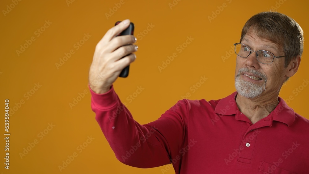 Happy smiling elderly bearded man 50s wears yellow shirt doing selfie shot on mobile phone post photo on social network isolated on yellow background studio portrait Happy smiling elderly bearded man 50s wears yellow shirt doing selfie shot on mobile phone post photo on social network isolated on yellow background studio portrait