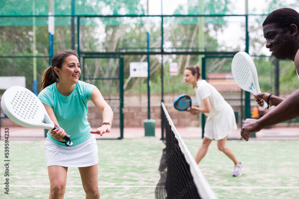 Emotional woman playing doubles paddle tennis with male and female opponents at warm sunny day, healthy lifestyle concept