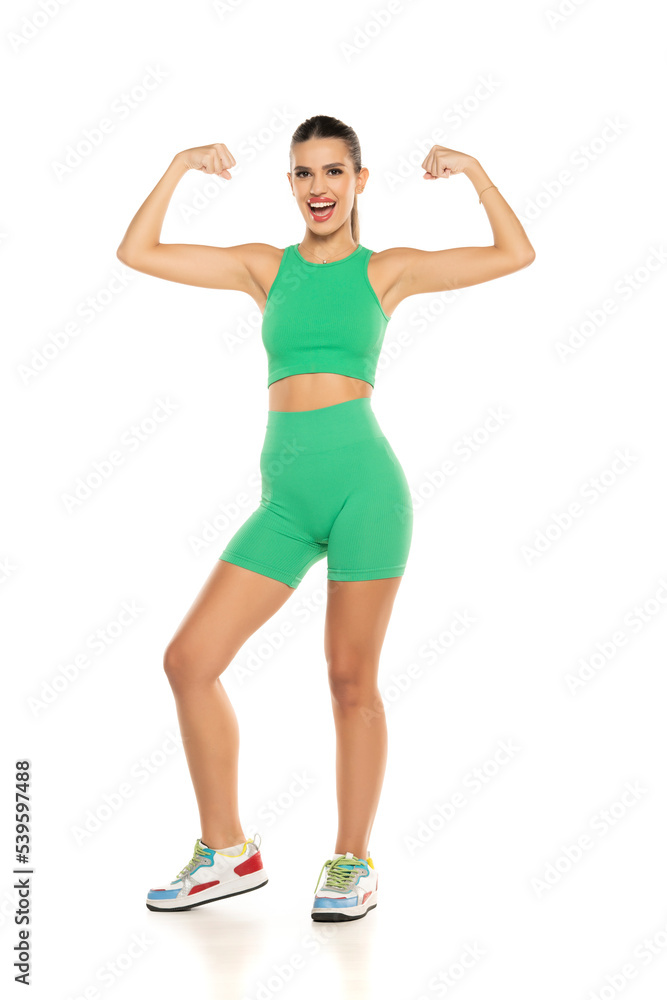 Fototapeta premium young sporty smiling woman in green shorts and top showing biceps on white background