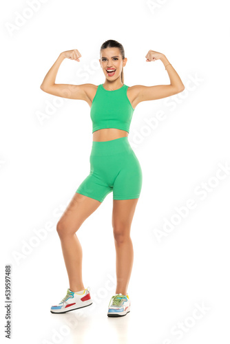 young sporty smiling woman in green shorts and top showing biceps on white background