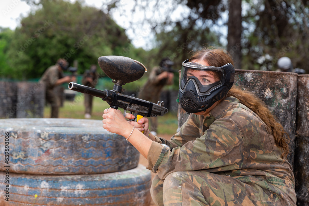 Fototapeta premium Female paintball player in protective mask with ammunition and gun on paintball field