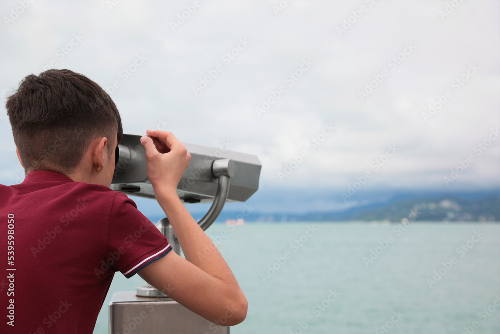 Teenage boy looking through mounted binoculars at mountains. Space for text