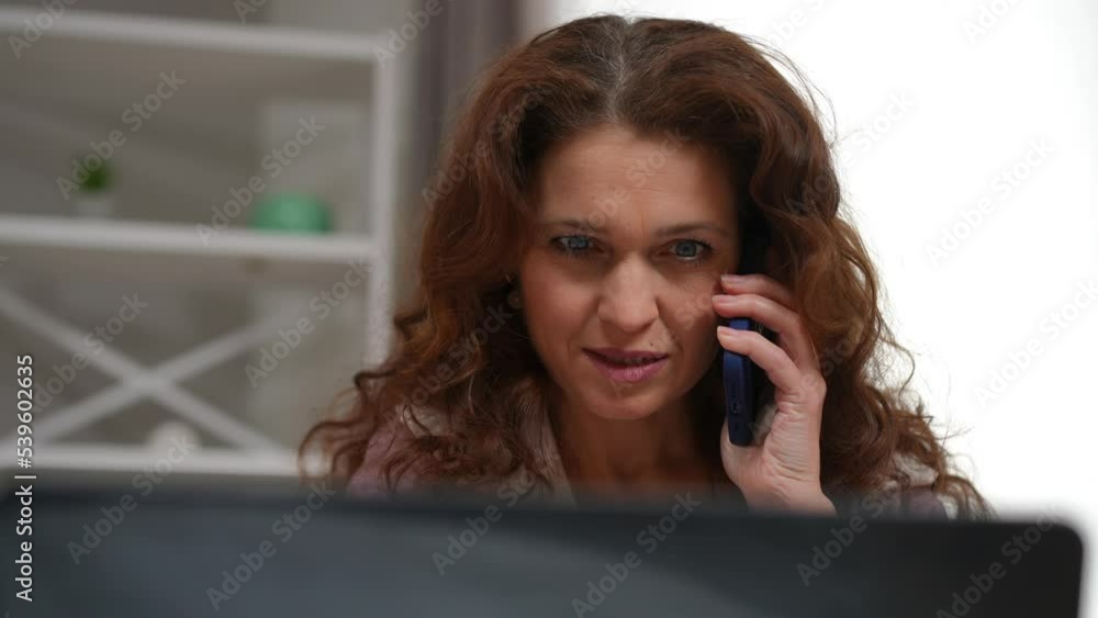 Close-up front view of confident positive female mature freelancer talking on phone surfing Internet on laptop. Beautiful Caucasian woman discussing business idea planning startup at home Close-up front view of confident positive female mature freelancer talking on phone surfing Internet on laptop. Beautiful Caucasian woman discussing business idea planning startup at home
