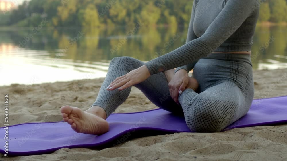 Unrecognizable athletic slender young woman folds her legs in lotus position helping herself with hands. girl in gray sports suit on purple fitness mat does yoga on bank of river or lake in park. Unrecognizable athletic slender young woman folds her legs in lotus position helping herself with hands. girl in gray sports suit on purple fitness mat does yoga on bank of river or lake in park.