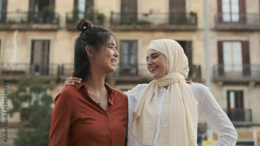 Young multiethnic female friends smiling and talking while hugging each other outdoors on the street. Young multiethnic female friends smiling and talking while hugging each other outdoors on the street.