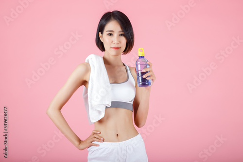 Portrait of smiling sporty woman with towel on her shoulder and holding drinking bottle of water on pastel pink background. © skarie