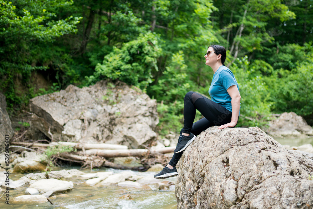 Obraz premium Woman hiker sitting on rock by river in the forest. Female exploring untamed natural areas along the river. Outdoor recreation, active leisure, local getaway concept