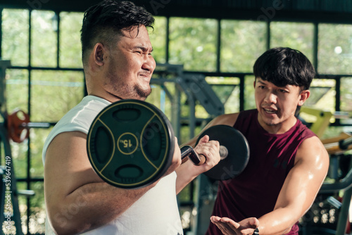 Plump asian man in loss weight course weight training is intensely lifting a barbell with his personal trainer giving an advise beside in the fitness centre, weight loose concept, healthy lifestyle.
