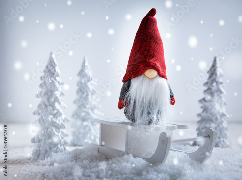 Cute Christmas gnome is sitting on sled, winter country with white trees photo