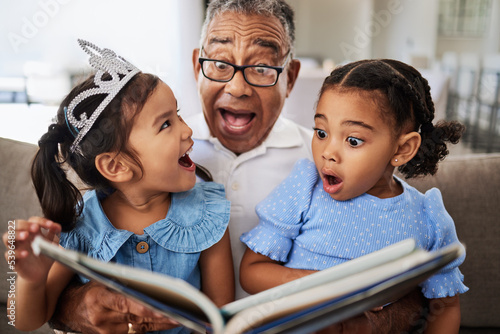 Family, reading book and children with grandpa for wow, surprised and excited expression in a living room with people bonding. Grandfather or man, kids and story time at home with man and girls photo