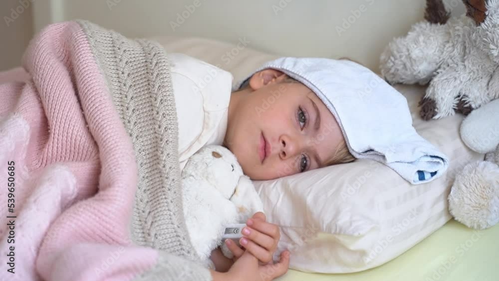 Sick fever cough child lying in bed with cold compress on his forehead, measure body temperature with electronic thermometer.Children cold and flu, coronavirus illness concept. Sick fever cough child lying in bed with cold compress on his forehead, measure body temperature with electronic thermometer.Children cold and flu, coronavirus illness concept.