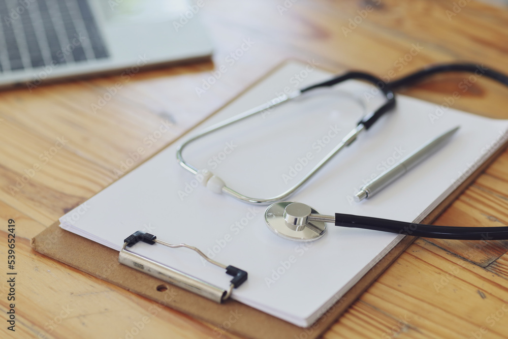 Stethoscope with prescription clipboard and Laptop and blood pressure meter on wooden table in office, close-up Stethoscope with prescription clipboard and Laptop and blood pressure meter on wooden table in office, close-up